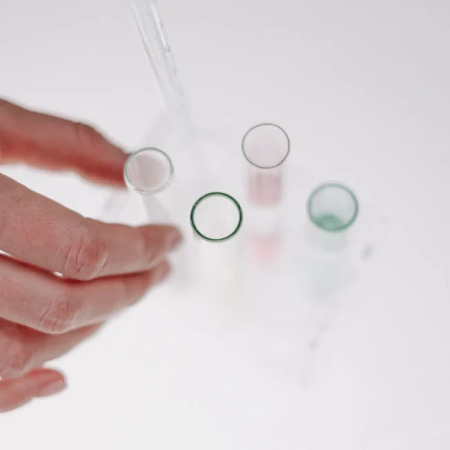 Dłoń sięgająca po szklaną probówkę ze stojaka na jasnym tle. Obok widoczna jest pipeta, co nawiązuje do laboratoryjnych badań i procesu tworzenia nowoczesnych formuł kosmetycznych.